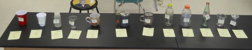 The twelve containers are labeled with sticky notes, while students' initial assessment of  thermal ranking is written on the paper pieces in front of the containers.